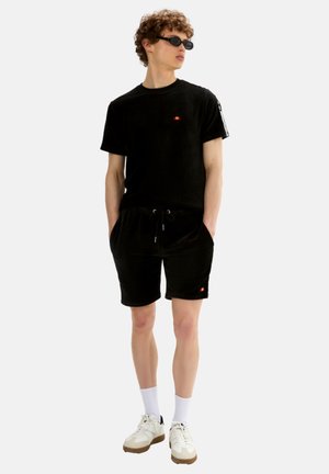 Joven con camiseta negra y pantalones cortos con logos, calcetines blancos, zapatillas y gafas de sol negras, de pie con las manos en los bolsillos.