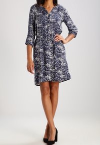 Marineblaues und weißes gemustertes Kleid mit Dreiviertelärmeln, V-Ausschnitt, taillierter Taille und schwingendem Rock; getragen mit schwarzen High Heels.