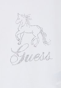 Άσπρο μπλουζάκι με σχέδιο ενός αλόγου από ασημί στρας πάνω από τη λέξη "Guess", επίσης αποδοσμένη με ασημί στρας.