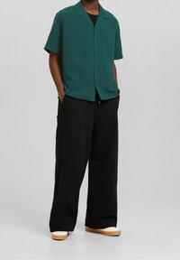 Tyrkysová texturovaná košile s krátkým rukávem a límečkem, zapínání na knoflíky, v kombinaci s černými kalhotami širokého střihu a bílou obuví s hnědými akcenty.