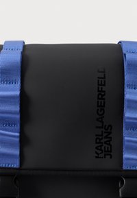 Juoda stačiakampė rankinė su „KARL LAGERFELD JEANS" juodu logotipu, pritvirtinta platiais mėlynais dirželiais su siūlėmis ir juodu plastikiniu sagtimi.