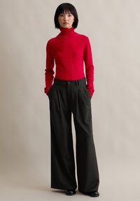 Suéter de cuello alto rojo brillante en punto acanalado, combinado con pantalones negros de pierna ancha de cintura alta que tienen plecos frontales y bolsillos laterales.