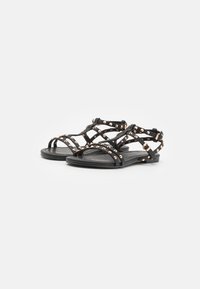 Svarta platta sandaler med korsade remmar, prydda med metallic-nitar. Har en slät sula och minimalistiskt ankelsupport.