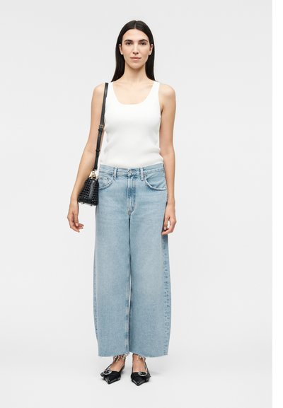 Νέα γυναίκα με μακριά σκούρα μαλλιά, φοράει λευκό μπουστάκι, φαρδιά ανοιχτό μπλε τζιν, μαύρα μυτερά παπούτσια και κρατά μια μαύρη τσάντα ώμου.