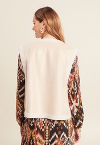 Cremfarbener ärmelloser Weste mit einem weichen, strukturierten Kragen. Das Modell trägt ein gemustertes, farbenfrohes Kleid mit filigranen Designs.