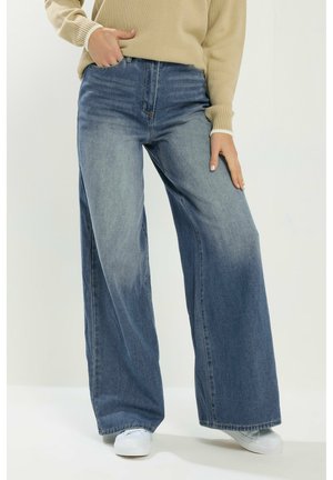 Persoon die een wijde blauwe spijkerbroek, een beige trui en witte sneakers draagt, met één hand in de zak en de andere rustend op de dij, staand op een witte vloer.