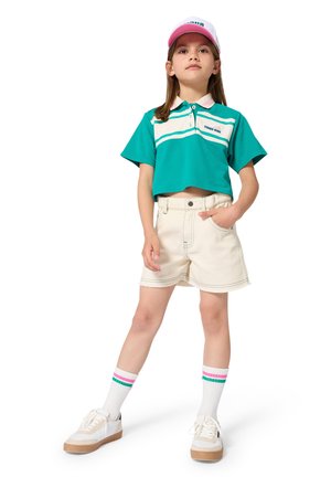 Młoda dziewczyna stoi pewnie, ubrana w turkusową, skróconą koszulkę polo, beżowe szorty, białe trampki, paskowane skarpetki i różowo-białą czapkę z daszkiem.