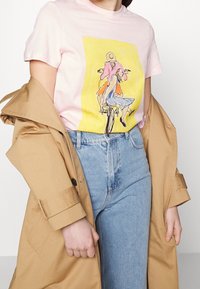 Camiseta de algodón rosa con una ilustración colorida de una bicicleta, combinada con unos jeans de mezclilla azul de cintura alta y un abrigo beige.