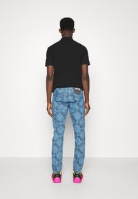 Schwarzes Poloshirt, hellblaue Jeans mit durchgehendem Logo-Muster und mehrfarbigen Turnschuhen. Die Jeans haben ein hinteres Markenetikett.