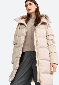 Beige gewatteerde jas met hoge kraag, met een ritssluiting aan de voorkant, gouden hardware en zijzakken. Draagt over een lichte trui en donkere broek.