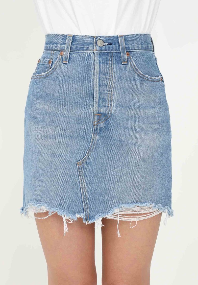 Mini-jupe en denim bleu clair avec ourlet effiloché et poches avant, portée par une personne devant un fond blanc uni.