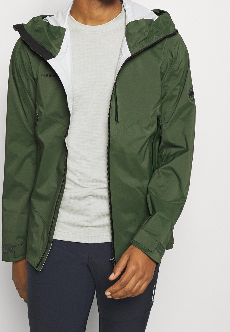 Zielona wodoodporna kurtka z kapturem wyłożonym białą podszewką, kieszeniami na zamek i regulowanymi mankietami, w połączeniu z jasnoszarą, teksturowaną koszulą.