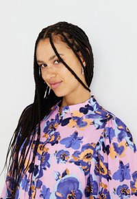 Blusa rosa con grande motivo floreale blu e arancione, design con bottoni e maniche a sbuffo. Tessuto morbido con una leggera lucentezza.