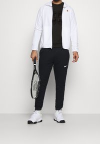 Biała kurtka na zamek, czarna koszulka z teksturowanym logo, czarne dresy z białym logo Nike i białe buty sportowe trzymające rakietę tenisową.