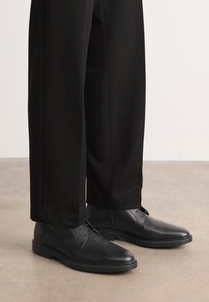 Schwarze Lederschuhe mit glatter Textur, runder Spitze und Schnürdesign, kombiniert mit schwarzen Hosen. Minimalistischer Stil mit niedrigem Absatz.