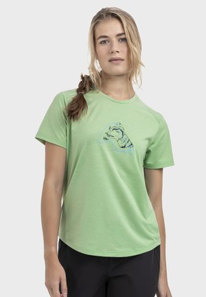 Junge Frau mit langem Zopf, die ein hellgrünes T-Shirt mit einem abstrakten schwarzen und blauen Muster auf der Brust trägt und vor einem einfarbigen Hintergrund steht.