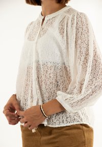 Blusa bianca in pizzo con collo alto, maniche lunghe a sbuffo e chiusura con bottoni. Tessuto leggero con motivi floreali e caduta morbida.