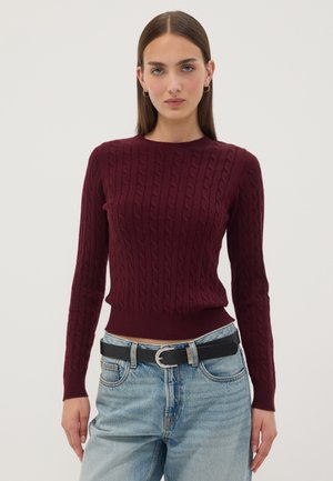 Donna con capelli castani lisci che indossa un maglione bordeaux a treccia, jeans azzurri chiari e una cintura nera con fibbia argentata.
