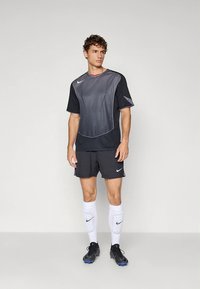 Musta ja harmaa urheiluasu, joka sisältää lyhythihaisen tekstuurikuvioidun paidan, siihen sopivat shortsit, valkoiset sukat ja mustat nappikengät.