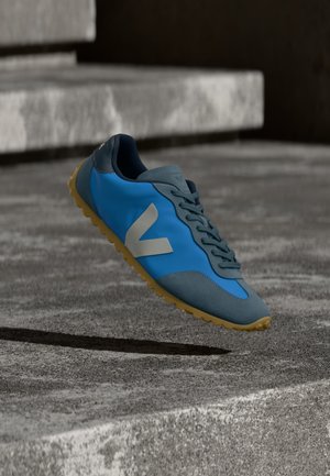 Blauer und grauer Sneaker mit Gummisohle und weißem "V"-Logo, schwebend über groben Betontreppen im Freien.