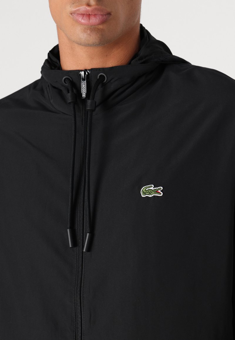 Veste à capuche noire en matériau léger, dotée d'une fermeture éclair centrale et d'un petit logo crocodile brodé en vert sur la poitrine.