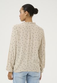 Beige blouse with black polka dots, long sleeves, ruffled neckline, textured fabric, rounded hem, worn with light blue jeans.