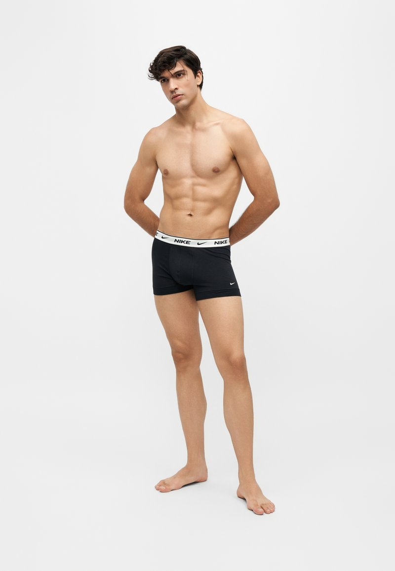 Homme torse nu aux cheveux foncés portant des boxers noirs Nike, debout pieds nus devant un fond blanc uni, les mains dans le dos.