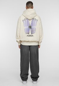 Hoodie beige avec un grand motif de papillon violet au dos, comportant un texte en dessous. Assorti à un pantalon gris ample et des baskets blanches.