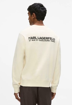 Mannelijk model met de rug naar de camera, draagt een crèmekleurige trui met de tekst "KARL LAGERFELD 21 RUE ST-GUILLAUME PARIS" in zwart op de achterkant gedrukt.