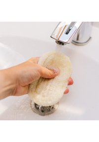 Loofah pad in a hand, under running water from a chrome faucet. The pad is textured, light-colored, and oval-shaped, with a thick edge.