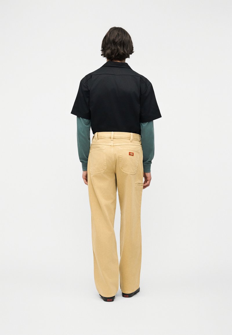 Persona con cabello corto y oscuro, vistiendo una camisa negra de manga corta sobre una camisa verde de manga larga y pantalones beige de pierna ancha, de pie de espaldas.