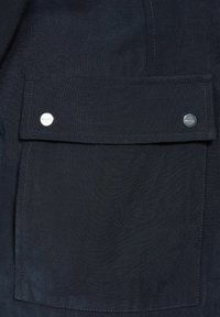 Marineblauem Stoff mit strukturierter Oberfläche, der eine Klapptasche enthält, die mit zwei silbernen Druckknöpfen gesichert ist und sichtbare Nähdetails aufweist.