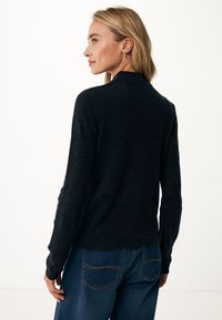 Tmavě šedý svetr z vlněné směsi s kulatým výstřihem, dlouhými rukávy a žebrovanými manžetami, v kombinaci s modrými džíny s oranžovým prošíváním.