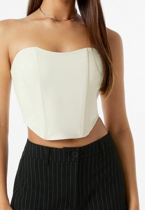 Kobieta nosząca kremowy gorset bez ramiączek w połączeniu z czarnymi, wysokim stanem spodniami w prążki oraz cienki srebrny naszyjnik typu choker.