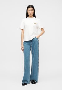 Γυναίκα που στέκεται με το χέρι στην τσέπη, φορώντας λευκό μπλουζάκι με μικρό λογότυπο και μπλε παντελόνι με φαρδιά πόδια και πλευρική ρίγα, καθώς και μαύρα παπούτσια.