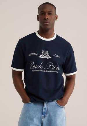 Jeune homme portant un t-shirt bleu marine avec bordure blanche et texte, associé à un jean bleu clair, debout les mains dans les poches.