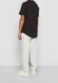 Mężczyzna w czarnej koszulce z krótkim rękawem, jasnych spodniach cargo i białych sneakersach marki Calvin Klein, stojący na tle gładkiego jasnoszarego tła.