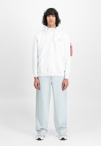 Λευκή φούτερ με καγκουρό τσέπη, κουκούλα με κορδόνι και λεπτομέρεια λογότυπου. Συνδυασμένη με ανοιχτό μπλε παντελόνι wide-leg και αθλητικά παπούτσια vintage.