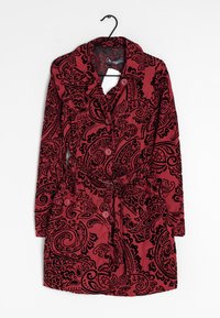 Rood ceintuurcoat met zwarte paisleypatronen, met knopen aan de voorkant, lange mouwen en twee klepzakken, hangend aan een zwarte hanger.