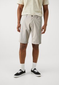 Shorts din bumbac bej, cu o croială lejeră și lungime până la genunchi, dotate cu închidere cu nasture, bucle pentru curea și buzunare laterale. Adidași negri cu șireturi albe.