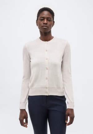 Γυναίκα που φοράει ένα ανοιχτού μπεζ καρό μπλουζάκι με κουμπιά και σκούρο ναυτικό παντελόνι, στέκεται μπροστά σε ένα απλό ανοιχτό γκρι φόντο.