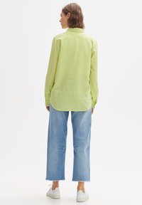 Woman standing facing away wearing a light green long-sleeve shirt, cropped light blue wide-leg jeans, and white sneakers on a white background.