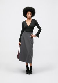 Vrouw met afro-kapsel in een zwarte lange mouwen top, grijze midi-rok, zwarte sokken, zwarte hakken en een kastanjebruine handtas in haar hand.