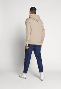 Sudadera con capucha beige con una capucha grande y suave, combinada con pantalones vaqueros oscuros y zapatillas blancas de caña alta. Tejido suave y diseño de ajuste relajado.