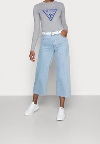 Osoba ubrana w szary długi rękaw z logo Guess, jasnoniebieskie szerokie dżinsy o kroju 7/8, biały pasek i białe trampki, stojąca na jednolitym tle.