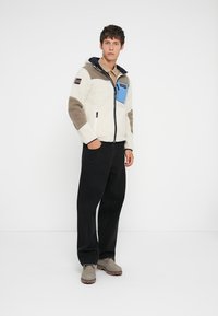 Man standing wearing a white and beige fleece jacket with a blue chest pocket, black pants, and gray boots against a white background.