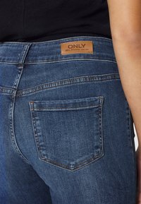 Jeans de mezclilla azul oscuro con un diseño ajustado, que presentan un parche de marca de cuero y bolsillos traseros con detalles de costura en contraste.