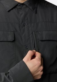 Camicia nera con bottoni realizzata in tessuto leggero, con due tasche sul petto e una tasca con zip sul lato sinistro, dotata di un colletto strutturato.