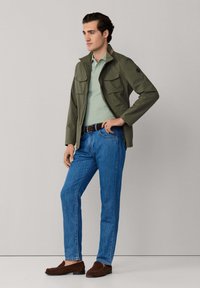 Muž v zelenej bunde, svetlozelenej polo košeli, modrých džínsoch, hnedom opasku a hnedých mokasínach, stojaci pred jednoduchým svetlosivým pozadím.