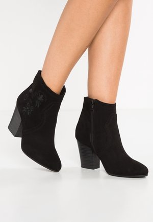 Zwarte suède enkellaarsjes met blokhakken, ritssluiting aan de zijkant en subtiele bloemrijke borduursels, gedragen op blote benen.